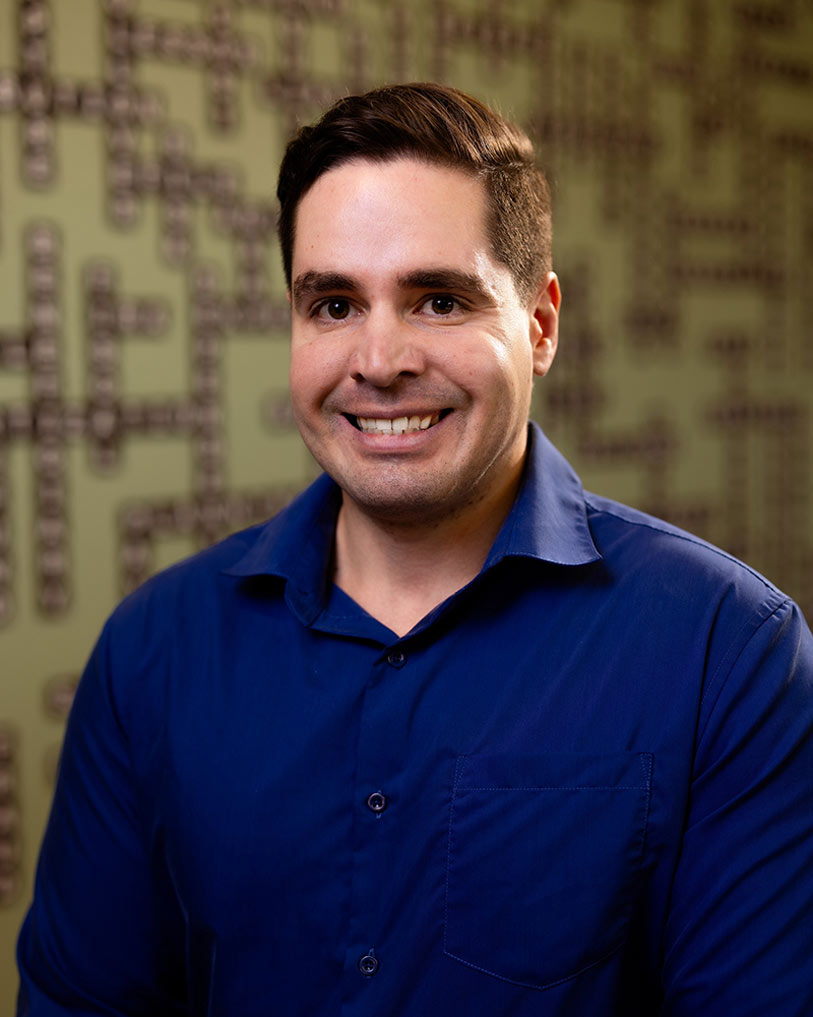 A man with short dark hair wearing a blue collared shirt smiles at the camera, standing in front of a green wall decorated with a crossword puzzle pattern.