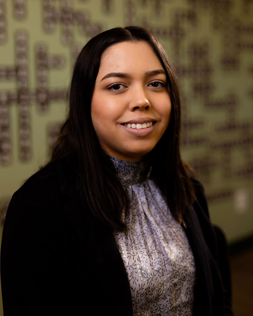 A young woman with long dark hair, wearing a patterned blouse and black jacket, smiles at the camera. The background is green with a geometric, textured design.