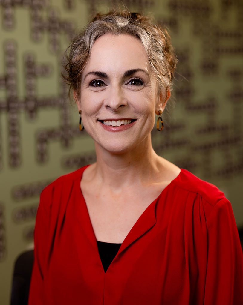 A woman with short, wavy gray hair, wearing a red blouse and earrings, smiles at the camera. The background is blurred with a green wall and dark patterns.
