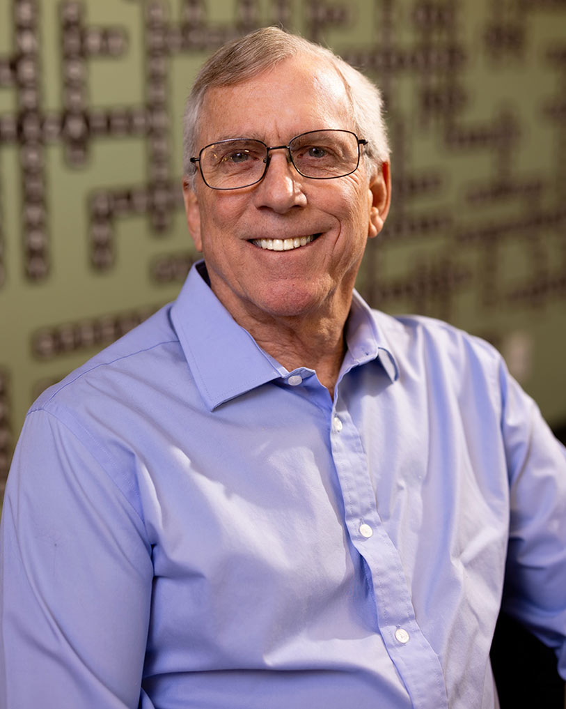 An older man with short gray hair and glasses is smiling at the camera. He is wearing a light blue button-up shirt and is seated in front of a green wall with a geometric pattern.