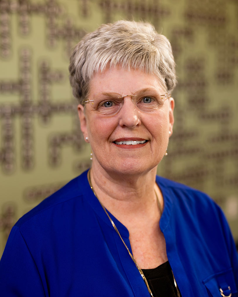 An older woman with short gray hair and glasses smiles at the camera. She is wearing a bright blue top and a necklace. The background is out of focus with a green wall and some dark patterns or text.