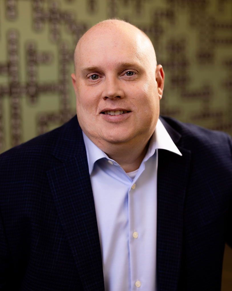 A bald man wearing a dark blue blazer and light blue shirt smiles at the camera. The background is softly blurred with a pattern of horizontal and vertical lines.