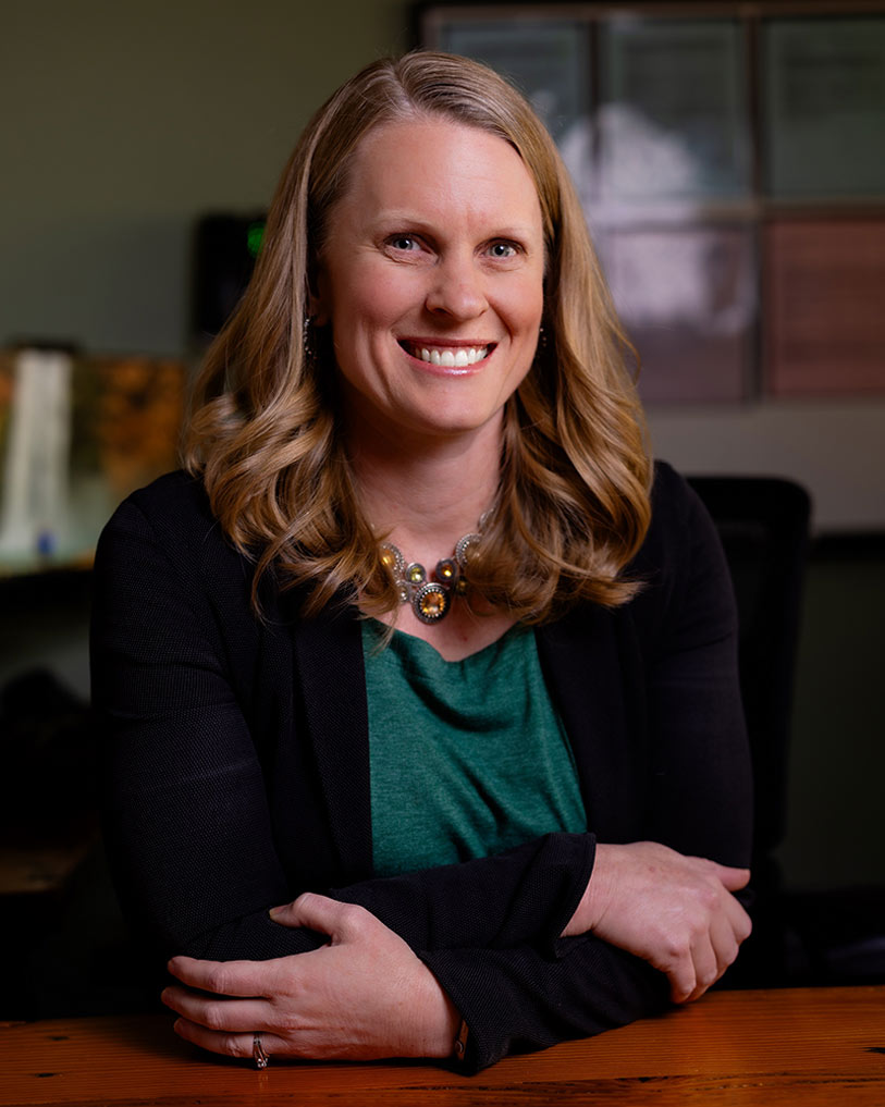 A smiling woman with wavy blonde hair sits at a wooden table, arms crossed. She wears a green top, black blazer, and a statement necklace, with framed documents blurred in the background.