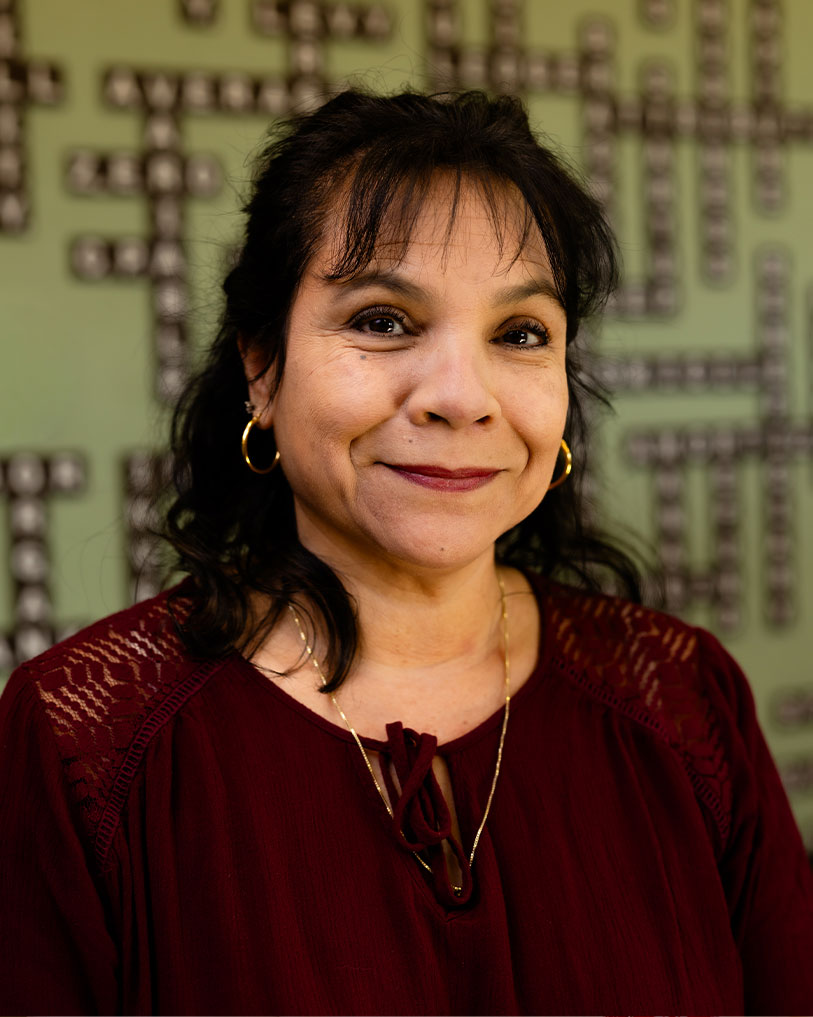 A woman with dark hair wearing gold hoop earrings and a burgundy blouse smiles at the camera, standing in front of a green wall with a crossword pattern.